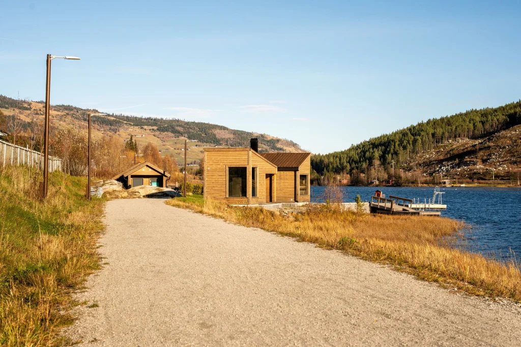 Grusvei foran i bildet. Badstova ca 100 meter unna. opplyst av solgult lys. Blå himmel og blått vatn.