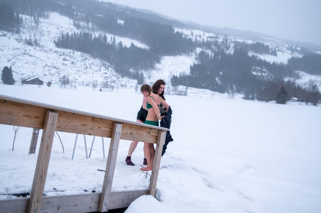Islagt vatn. Snø i lufta. To kvinner står i badetøy klare til å gå ned badestige til hol i isen der dei kan bade.